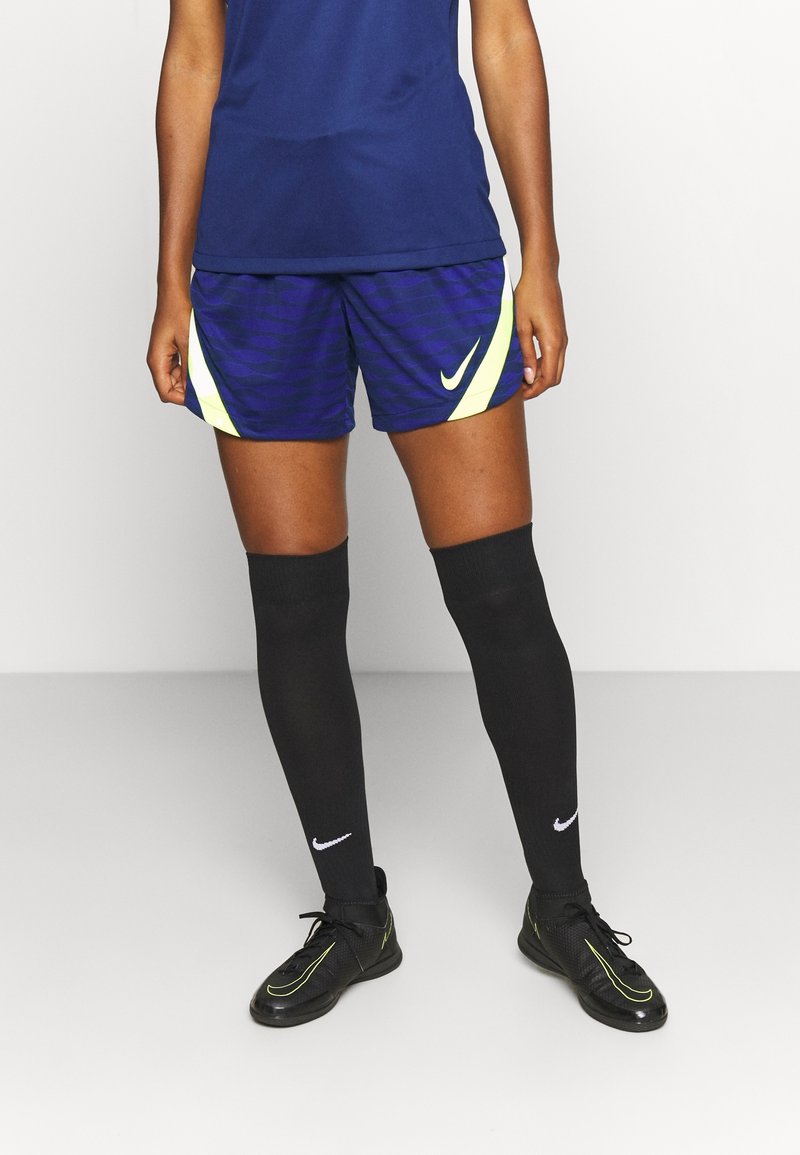 Nike urheilushortsit tummansinisinä, joissa on aaltoileva kuvio ja keltaisia yksityiskohtia. Yhdistetty mustiin polven yli ulottuviin sukkiin ja mustiin urheilukengiin.