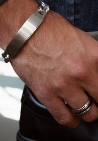 Zilveren armband met een vlakke, geborstelde metalen oppervlakte en in elkaar grijpende schakels; gedragen om een hand met zichtbare haren en ringen.