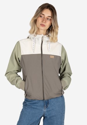 Woman wearing a color-block zip-up hooded jacket with green sleeves, white upper chest, gray body, and blue jeans, standing with hands in pockets.