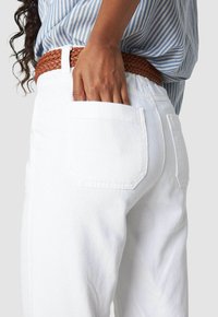 Mano con tono di pelle scura che riposa nella tasca posteriore di jeans bianchi indossati con una cintura intrecciata marrone e una camicia a righe blu.