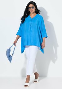 Ampio top blu brillante con scollatura a V, pantaloni bianchi aderenti e tacchi bianchi aperti. Gli accessori includono una borsa blu scintillante e occhiali da sole.