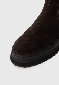 Botín de ante marrón hasta el tobillo con punta redondeada, costuras en contraste y suela de goma negra texturizada. El diseño es elegante y minimalista.