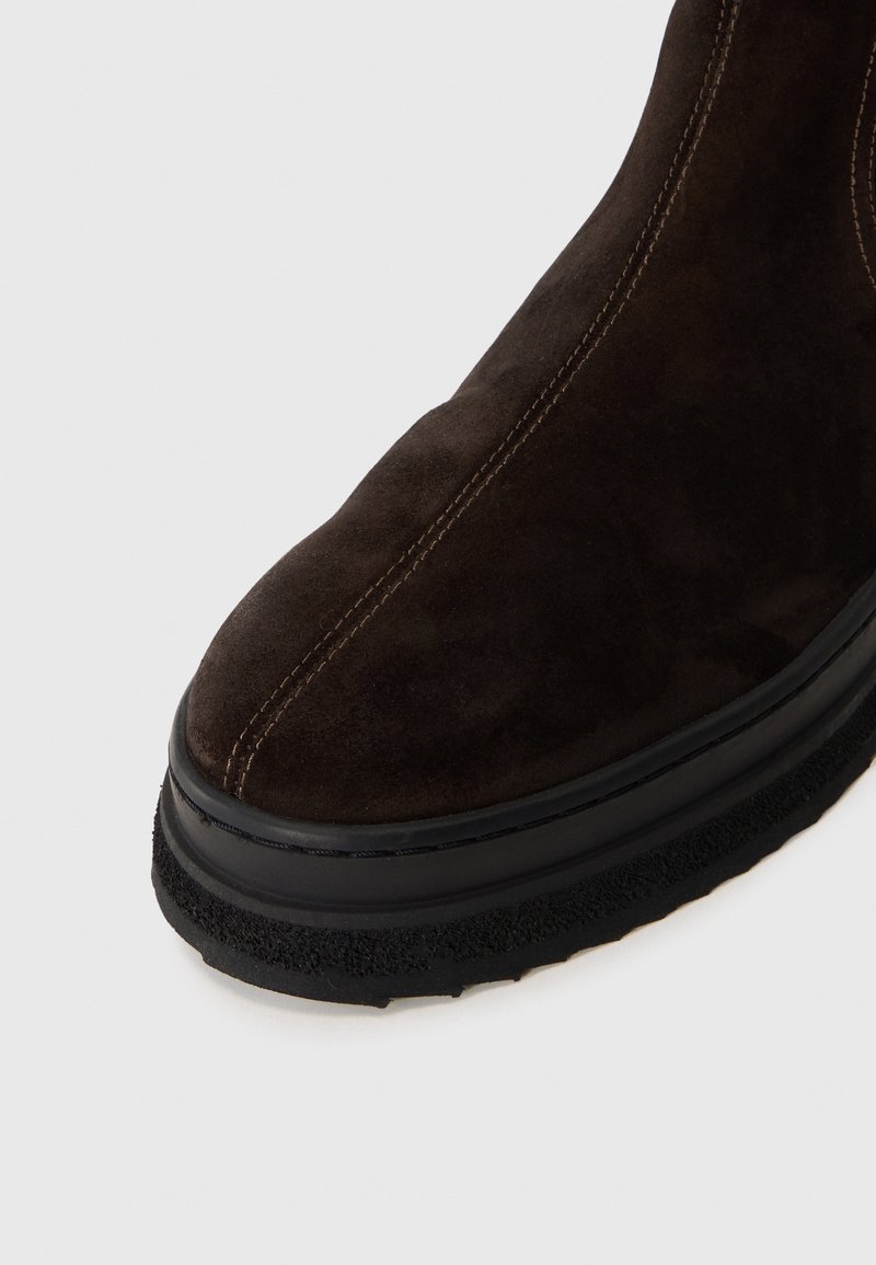 Stivale alla caviglia in suede marrone con punta arrotondata, cuciture a contrasto e suola in gomma nera testurizzata. Il design è elegante e minimalista.