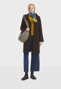 Cappotto di lana marrone con tasche, maglione a maglia giallo ocra, pantaloni a gamba larga blu, sciarpa con motivo, borsa grigia e stivali ankle marroni.