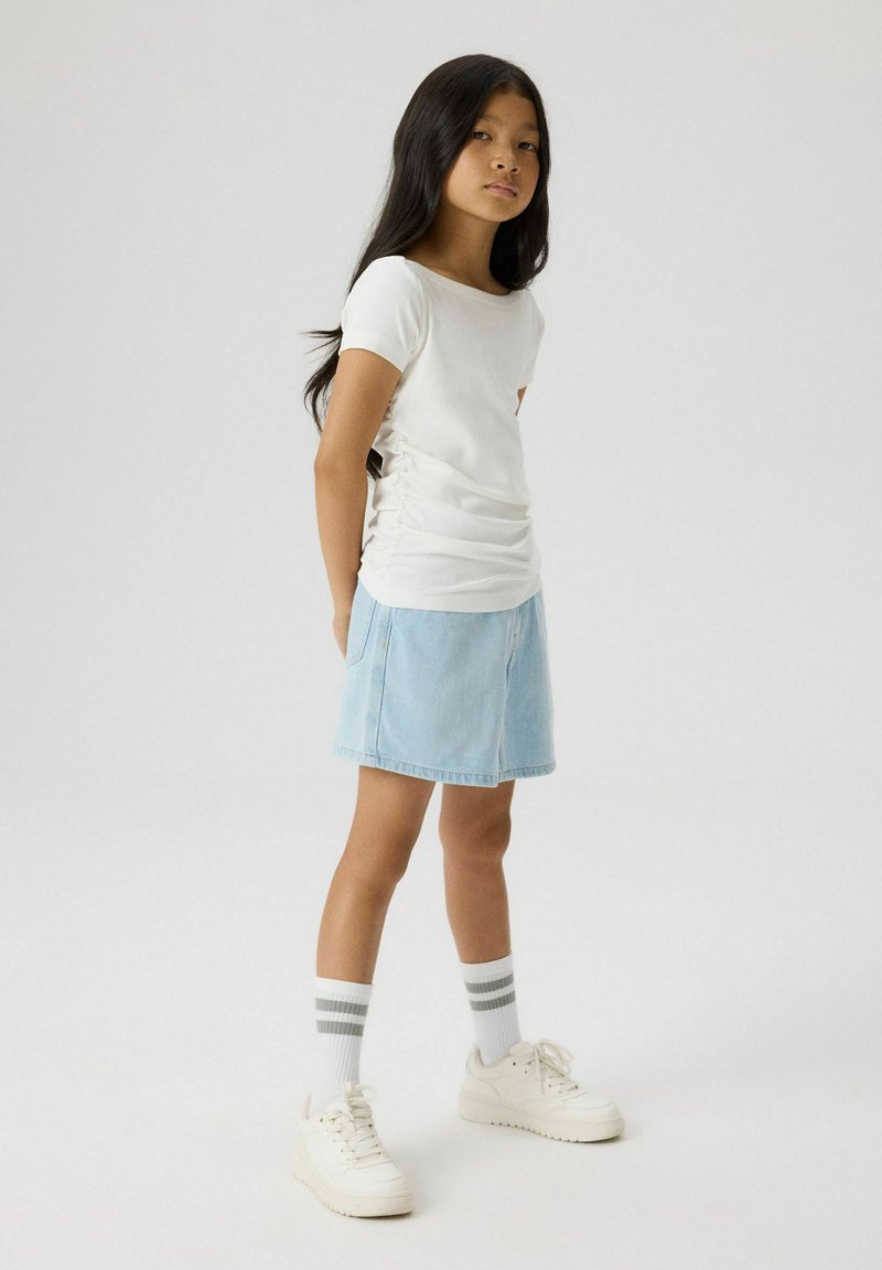 Fille aux longs cheveux foncés debout portant un haut blanc à manches courtes, une jupe en jean bleu clair, des baskets blanches et des chaussettes blanches avec deux rayures grises.