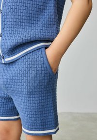 Plavi pleteni dvoslojni set s teksturiranim uzorkom, kratkim rukavima i bijelim prugastim detaljima na kratkim hlačama i izrezu.