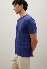 Junger Mann in einem blauen kurzärmeligen T-Shirt und beigen Hosen steht im Profil vor einem neutralen hellen Hintergrund mit den Händen hinter dem Rücken.