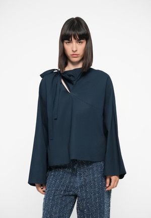 Navy blue blouse with a bow tie detail, asymmetric neckline, and long bell sleeves, made of smooth fabric. Paired with textured blue pants.