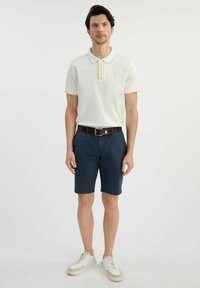 Biała koszulka polo z trzema guzikami, z żółtymi i niebieskimi paskami, w połączeniu z granatowymi szortami i białymi trampkami. Zawiera brązowy pasek.