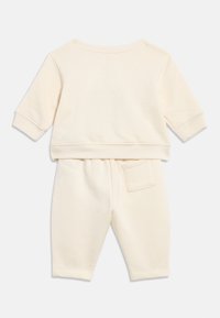Babykleidung aus weichem, cremefarbenem Stoff mit langen Ärmeln, Hose mit elastischem Bund und einer kleinen Tasche auf der Rückseite rechts.