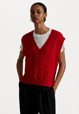 Rødt kabelstrikket ermeløst genser med dyp V-hals og ribbestrikket hem, brukt over en hvit kortermet T-skjorte. Minimalistisk design.