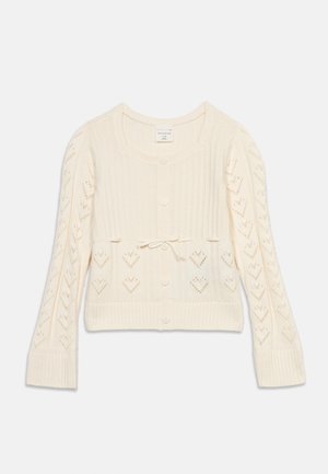Otroški kremasto pleteni jopič z odprtim vzorcem srčkov na rokavih, rebrasto teksturo, gumbi na sprednji strani in zavezanim lokom v pasu.