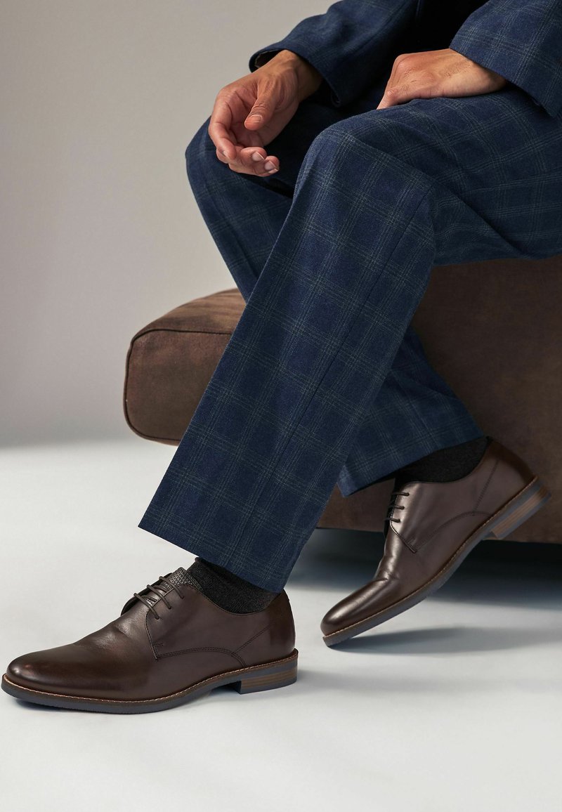 Braune Lederdressschuhe mit glatter Oberfläche, Schnürdesign und niedrigem Holzabsatz. Getragen mit navy-karierten Anzughosen und dunklen Socken.