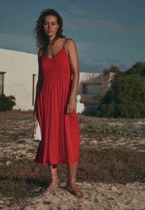 Naine seisab väljas, kandmas punast varrukateta kleiti ja valgeid sandaale, hoides valget lillekirjutusega käekotti.