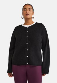 Cardigan noir texturé avec un col rond, comportant des boutons blancs sur le devant. Porté sur un haut blanc avec un col visible.