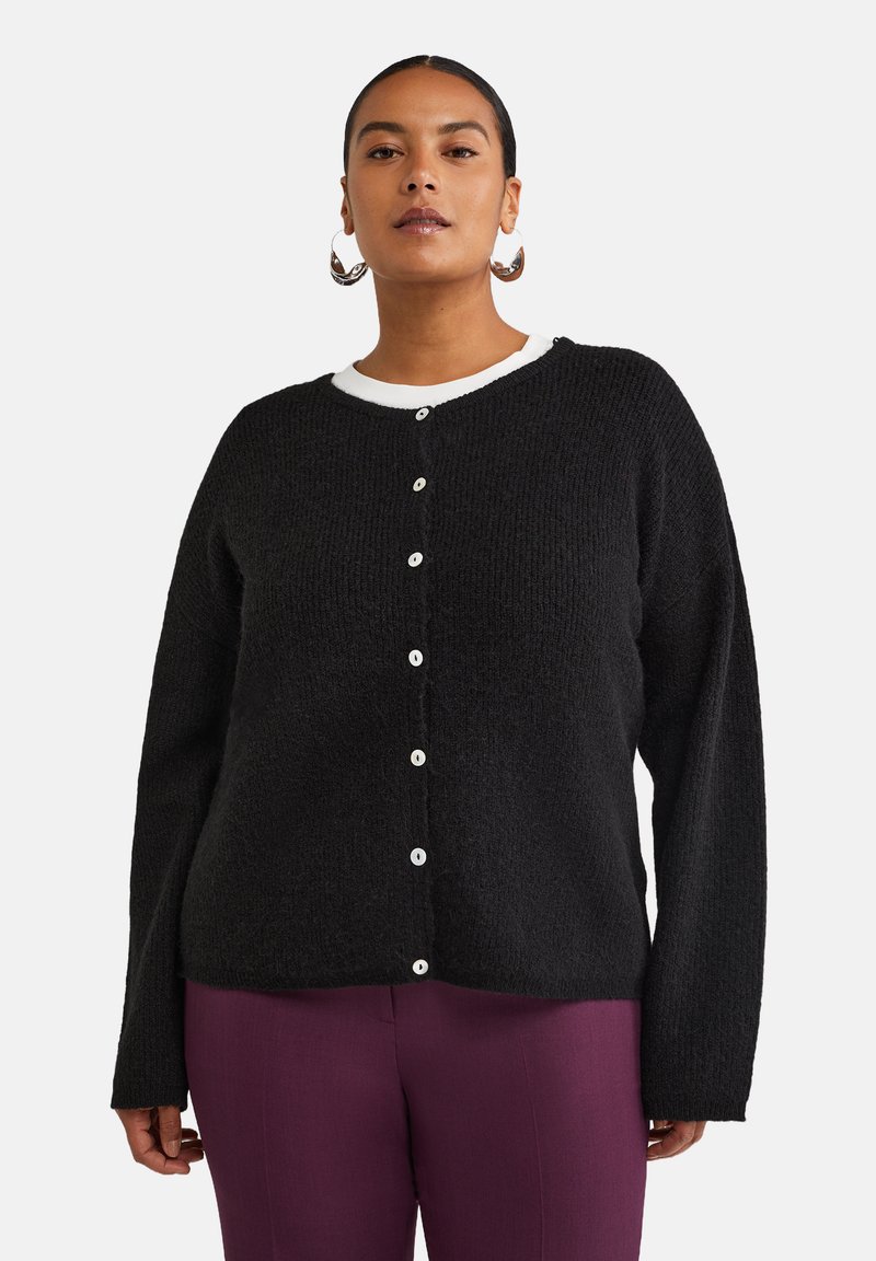 Cardigan noir texturé avec un col rond, comportant des boutons blancs sur le devant. Porté sur un haut blanc avec un col visible.