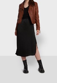 Braune Lederbikerjacke, schwarzes Strickoberteil, schwarze knielange Rock mit seitlichen Schlitz, und schwarze klobige Stiefeletten, vor einem neutralen Hintergrund.