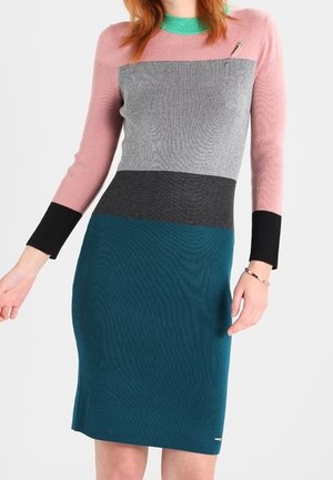 Femme portant une robe en tricot à blocs de couleurs avec des manches roses, un col vert, un buste gris, une taille noire et une jupe bleu sarcelle, debout devant un fond blanc.