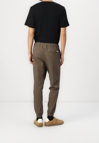 Osoba stoi tyłem, ubrana w czarną koszulkę, brązowe spodnie dresowe na elastycznej talii, czarne skarpetki i beżowe sneakersy z białymi akcentami na pięcie.