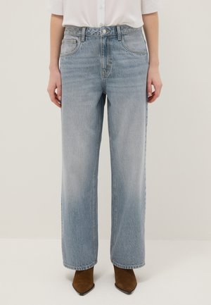 Persona con vaqueros de pierna recta azul claro, camisa blanca metida por dentro y botas marrones de punta afilada, de pie frente a un fondo liso.