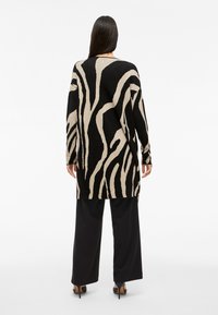 Γυναίκα με μακριά σκούρα μαλλιά, στραμμένη προς τα πίσω, φοράει ένα μαύρο και μπεζ μακρύ πουλόβερ με αφηρημένο σχέδιο, μαύρα φαρδιά παντελόνια και μαύρες γόβες.