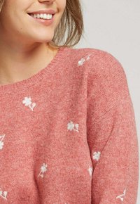 Pull en tricot rose avec des broderies florales blanches, encolure ronde et texture douce, mettant en avant un design décontracté avec des détails délicats.