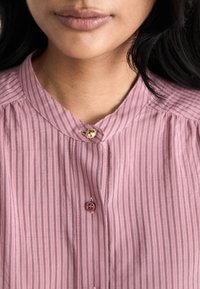Ροζ ριγέ μπλούζα από ελαφρύ ύφασμα. Διαθέτει όρθιο γιακά, κλείσιμο με κουμπιά και διακοσμητικό κουμπί στον λαιμό.