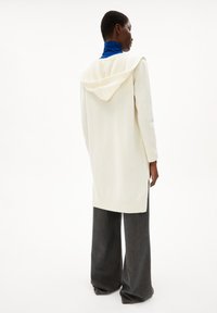 Langer cremefarbener Cardigan mit Kapuze, aus weichem Material, mit seitlichen Schlitzen und gerippten Bündchen, kombiniert mit dunklen, weit geschnittenen Hosen.