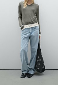 Maglietta grigia a maniche lunghe indossata sopra una camicia bianca, jeans azzurri chiari larghi, borsa nera con dettagli in borchie e scarpe nere flat.