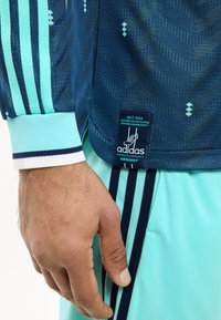 Εγγύς πλάνο ενός χεριού δίπλα σε σκούρα μπλε φανέλα της Adidas Γερμανίας με ετικέτα και ανοιχτό μπλε σορτς με σκούρες μπλε ρίγες.