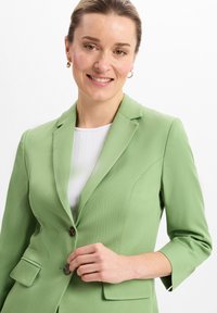 Hellgrüne Blazer mit taillierter Passform, Dreiviertelärmeln, Reverskragen und zwei Fronttaschen. Braune Knöpfe zieren die Vorderseite.