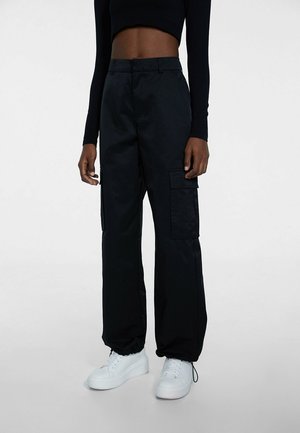 Personne portant un pantalon cargo noir avec poches latérales, un haut cropped noir à manches longues et des baskets blanches à plateforme, debout sur un fond gris.