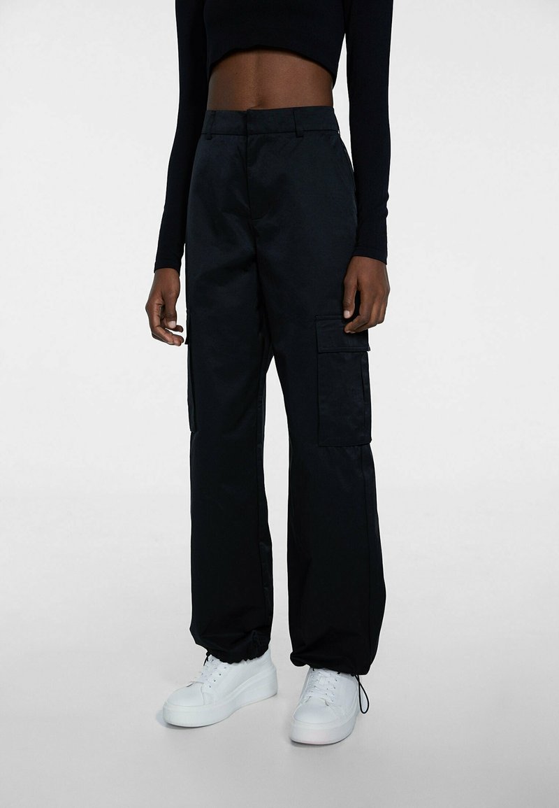 Personne portant un pantalon cargo noir avec poches latérales, un haut cropped noir à manches longues et des baskets blanches à plateforme, debout sur un fond gris.