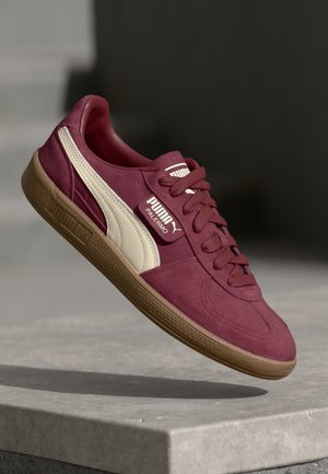 Burgundy sneaker with suede upper, cream accents, and lace closure. Features a rubber sole and a logo tag on the side.