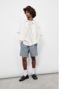 Modell som har på seg en løs hvit skjorte med knapper, grå shorts med snor, svarte sko, og hvite sokker. Sparsommelig bakgrunn.