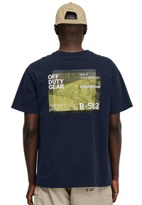 Navy blauwe katoenen t-shirt met een grote grafische print op de achterkant met de tekst "OFF DUTY GEAR" en verschillende nummers in wit en geel.
