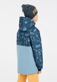 Giacca invernale blu e azzurro chiaro con cappuccio, caratterizzata da motivi geometrici, polsini elastici e una superficie liscia. Indossata con un berretto scuro.