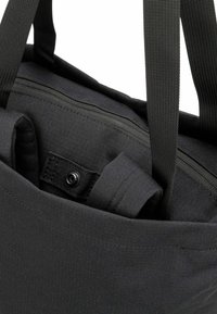 Bolso de mano negro con una tela texturizada, que cuenta con una parte superior con cremallera, asas resistentes y un botón a presión interno para el cierre.