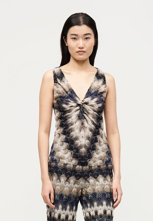 Γυναίκα με μακριά σκούρα μαλλιά που φοράει αμάνικη μπλούζα με V λαιμόκοψη και αντίστοιχο παντελόνι με επαναλαμβανόμενο κυματιστό ζιγκ-ζαγκ μοτίβο σε καφέ, μαύρο, μπλε και άσπρο.