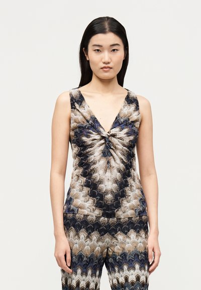 Femme aux longs cheveux foncés portant un haut sans manches à col en V et un pantalon assorti avec un motif zigzag ondulé répétitif en marron, noir, bleu et blanc.
