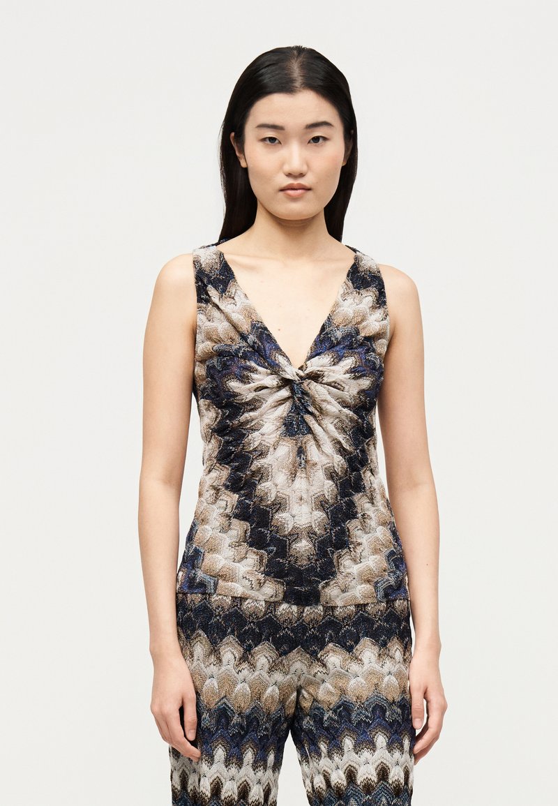 Γυναίκα με μακριά σκούρα μαλλιά που φοράει αμάνικη μπλούζα με V λαιμόκοψη και αντίστοιχο παντελόνι με επαναλαμβανόμενο κυματιστό ζιγκ-ζαγκ μοτίβο σε καφέ, μαύρο, μπλε και άσπρο.