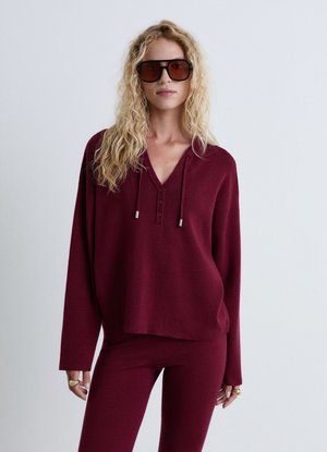 Blonde woman with curly hair wearing large dark sunglasses and a matching maroon long-sleeve top and pants set against a plain background.