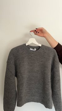 Sivi pleteni pulover z okroglim izrezom, spuščenimi rameni in rebrastim robom, obešen na beli obešalnik na svetlem ozadju.