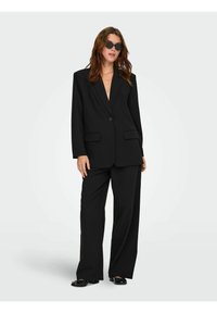 Sort oversized blazer med en enkelt knap, klapkrave og frontlommer, parret med vide bukser og sorte flade sko.