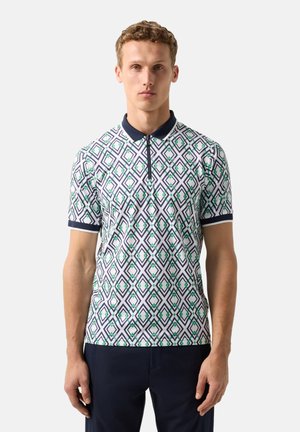 Mand iført kortærmet polo-shirt med marineblå krave og geometrisk diamantmønster i marineblå og grøn, kombineret med mørke bukser, stående mod en hvid baggrund.