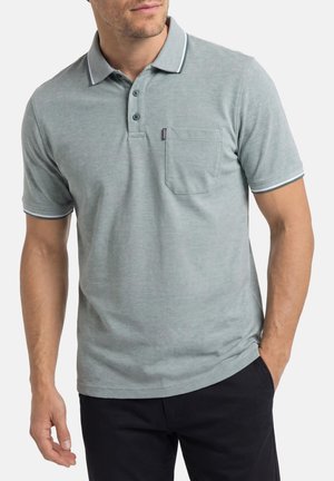 Man draagt lichtgrijs poloshirt met korte mouwen, drie knopen en een borstzakje, gecombineerd met donkere broek, staand met één hand in de zak.