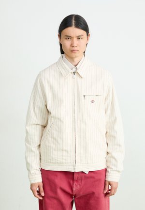 White striped jacket with a zip closure and chest pocket. Made of textured fabric, features a collar and ribbed cuffs. Worn over pink pants.