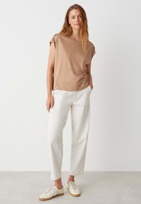 Tan t-shirt met korte mouwen en een ronde hals, gecombineerd met witte, gestructureerde, wijde broek en lichte sneakers op een neutrale achtergrond.