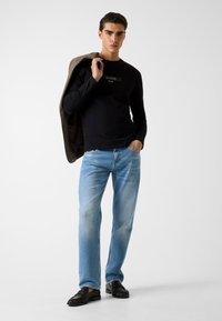 Zwart shirt met lange mouwen en het "GUESS"-logo, lichtblauwe spijkerbroek en zwarte leren loafers. Model draagt een bruine blazer over zijn schouder.