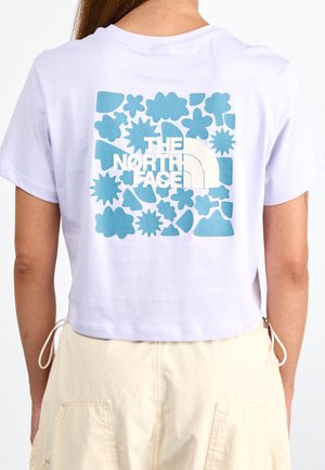 Osoba nosząca białą krótką koszulkę z niebieskimi kwiatowymi i abstrakcyjnymi wzorami oraz logo "THE NORTH FACE" na plecach, w połączeniu z beżowymi spodniami.